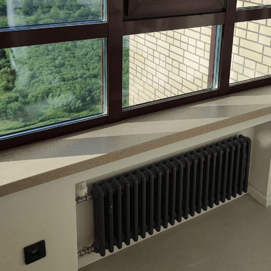 Светло-бежевый подоконник из акрилового камня в эркерной зоне с панорамными окнами и видом на зелёный лес, под подоконником — чёрная батарея