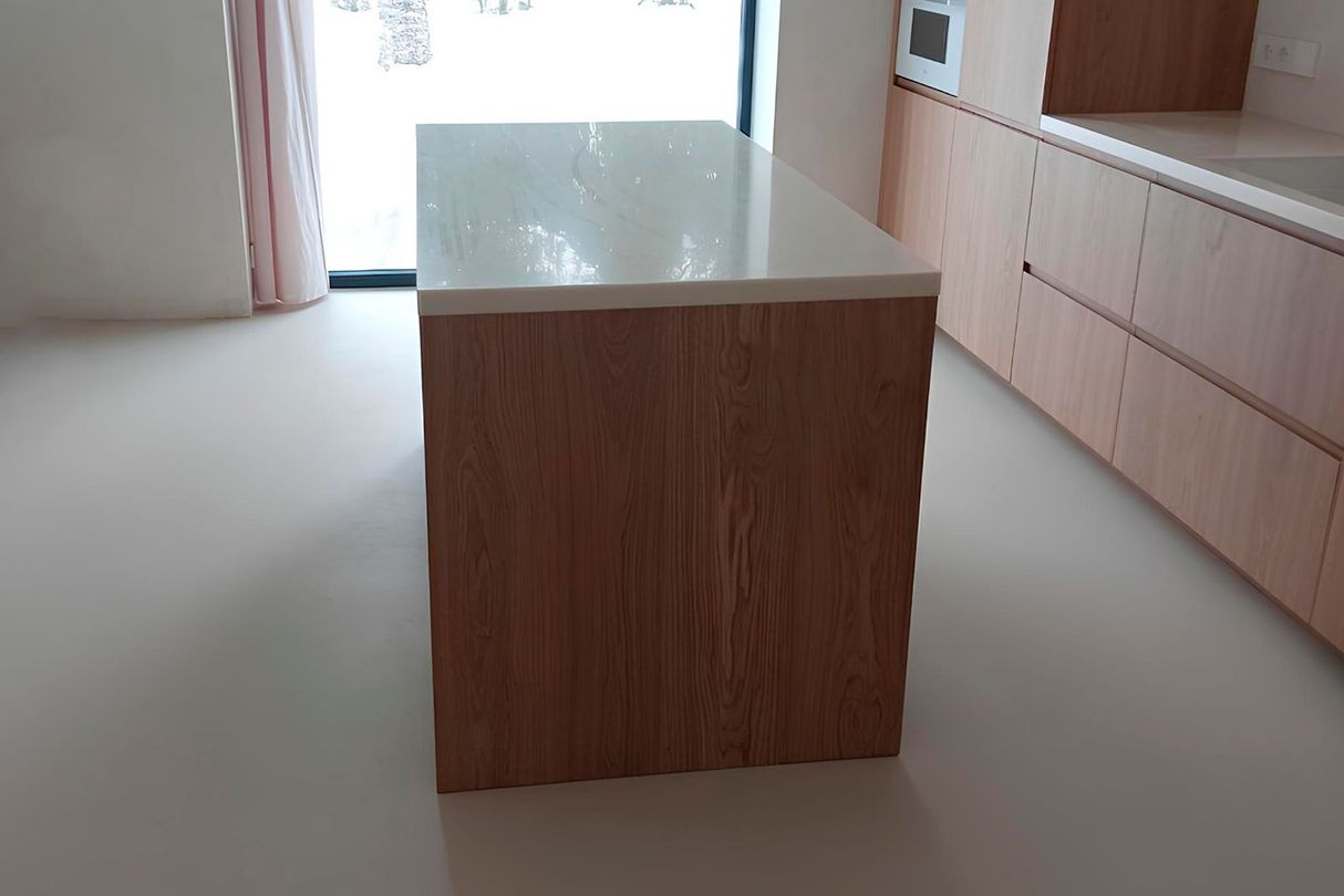 Кухонный остров с белой акриловой столешницей и фасадами под дерево в интерьере с панорамным окном и встроенной техникой.