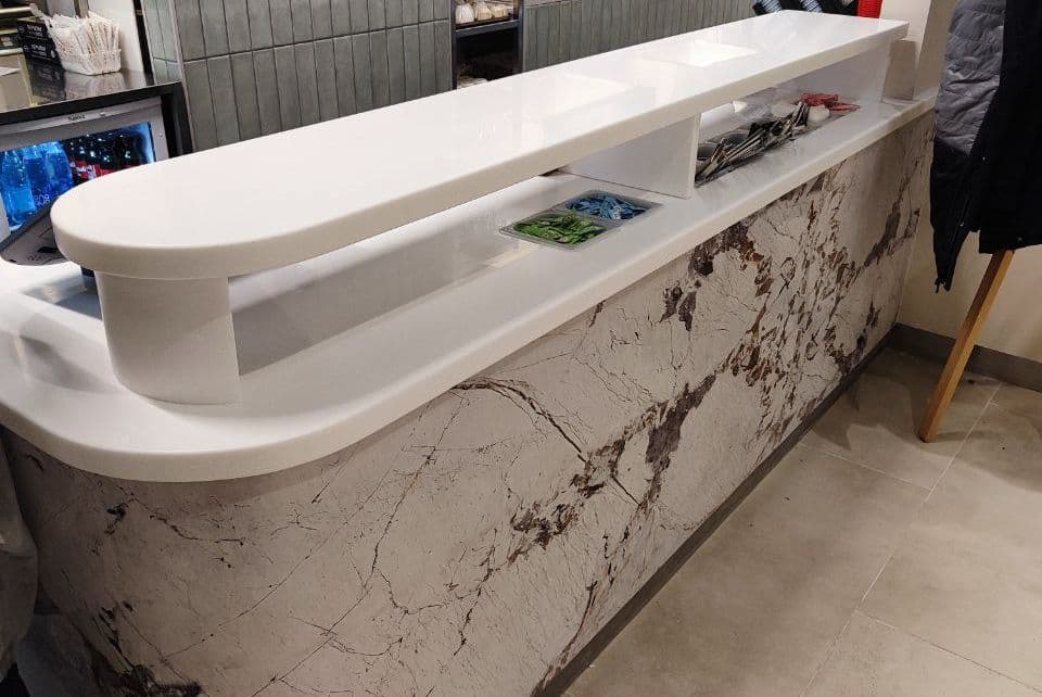 Белая глянцевая стойка из акрилового камня с верхней полкой и мраморным основанием, в современном интерьере кафе или ресторана.