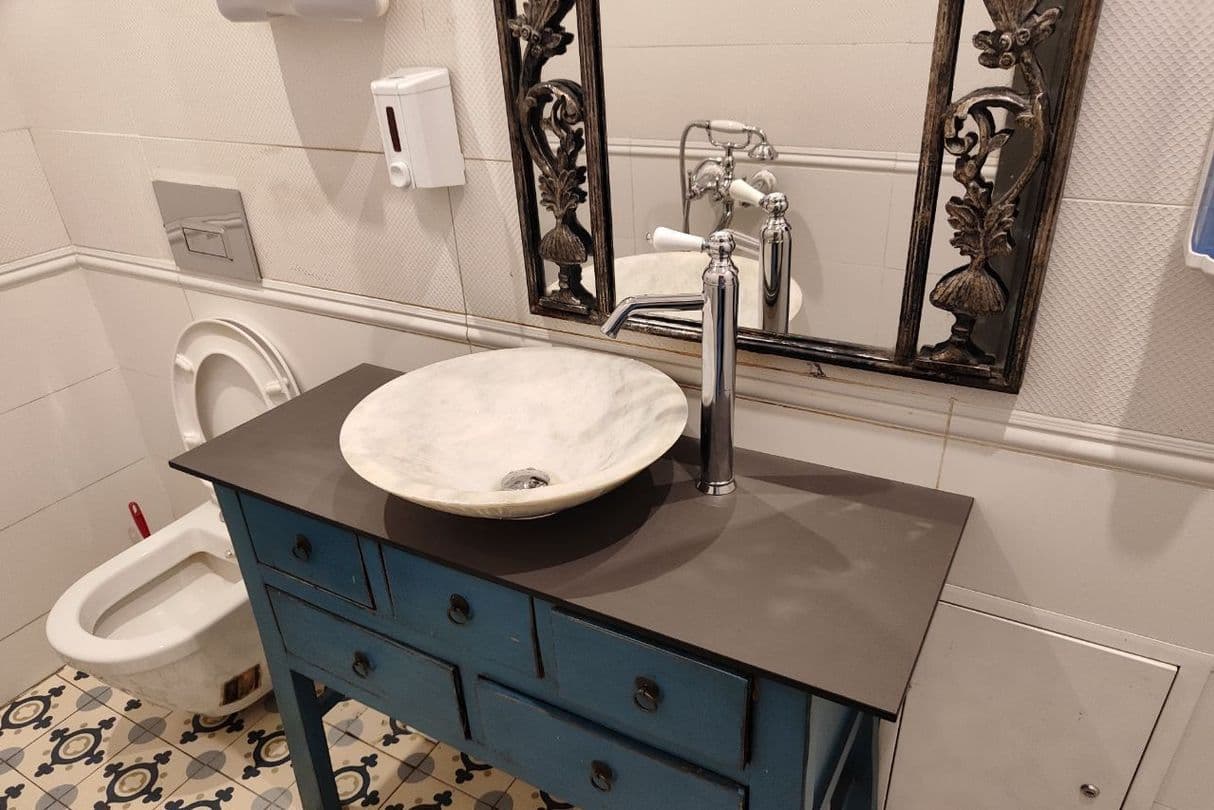 Темная столешница из HPL компакт пластика с накладной круглой раковиной из камня, установленная на синей тумбе в ванной комнате в винтажном стиле.
