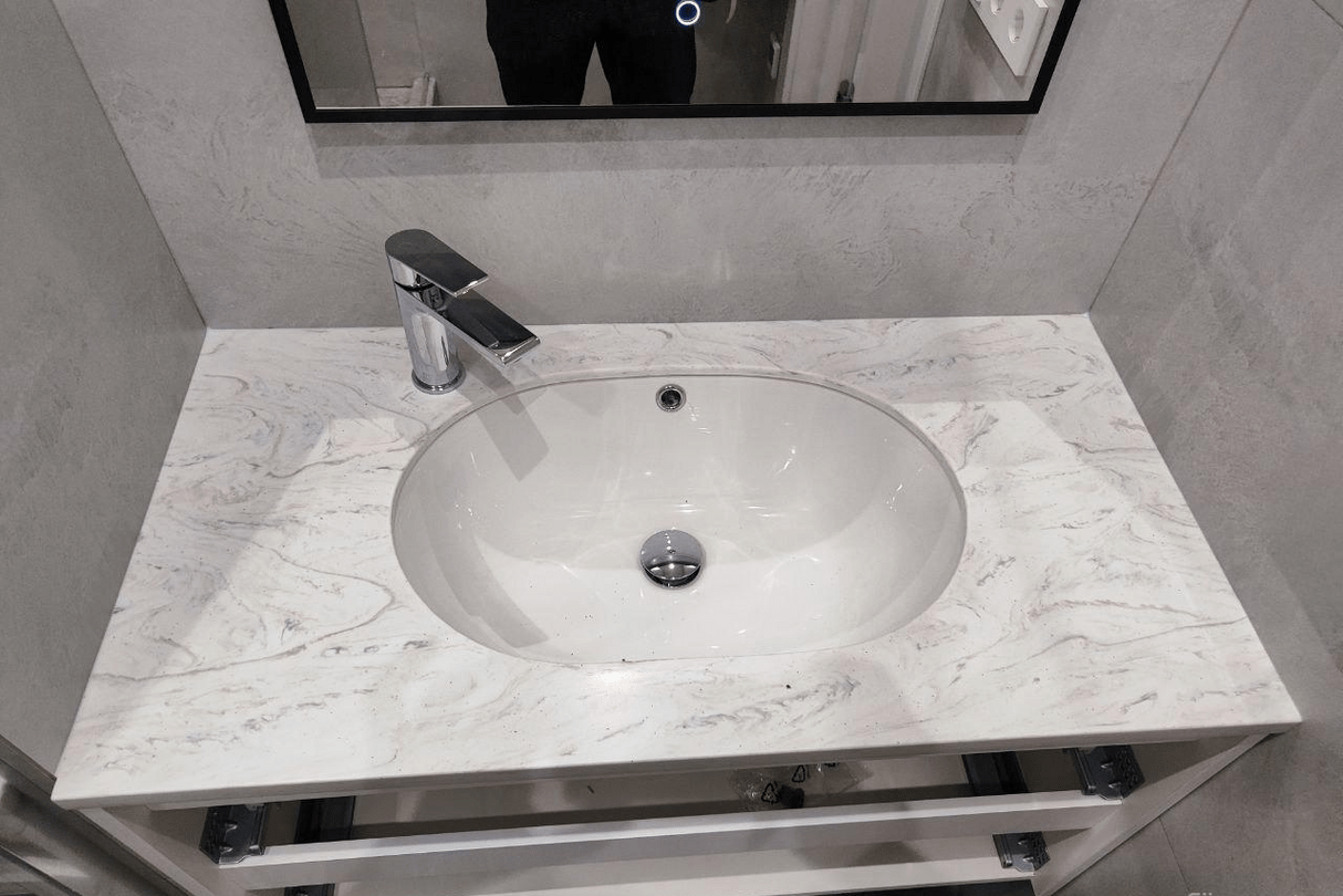 Раковина, встроенная в столешницу из светлого акрилового камня с мраморной текстурой, установленную в ванной комнате.
